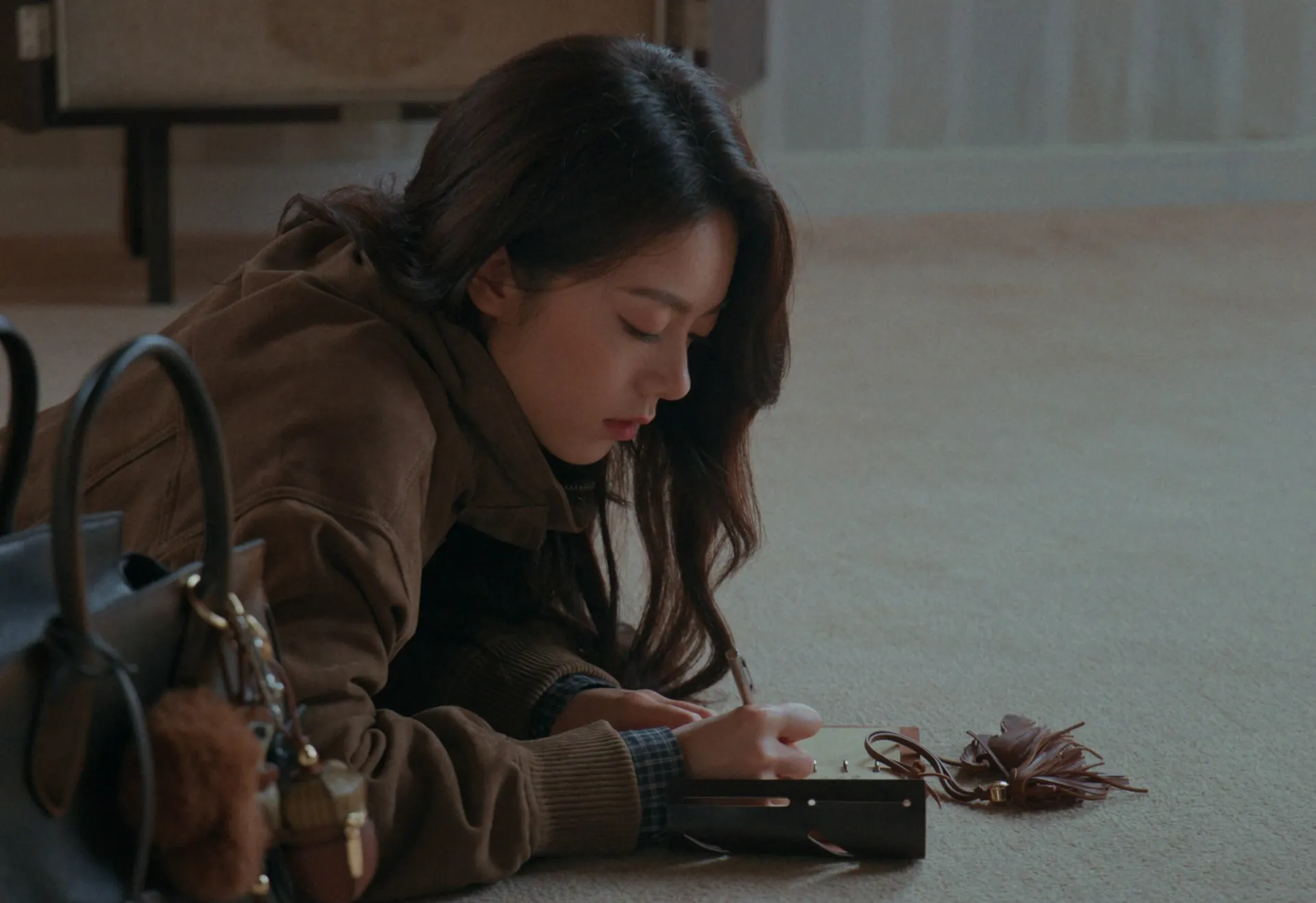 A woman in a suede jacket lying on a carpet, writing in a notebook beside a leather handbag